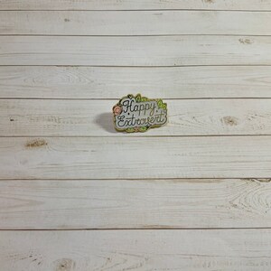 May include: A gold-colored enamel pin with a white background and green and pink floral accents. The pin says "Happy Extrovert" in white cursive lettering.