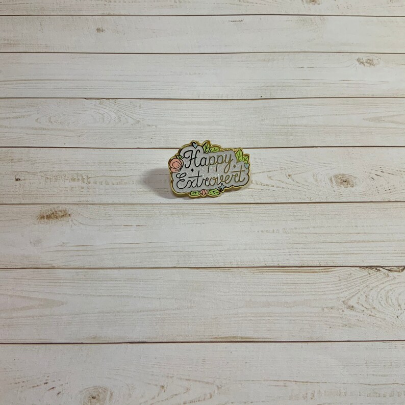 May include: A gold-colored enamel pin with a white background and green and pink floral accents. The pin says "Happy Extrovert" in white cursive lettering.