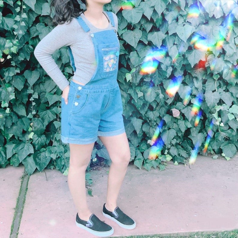 May include: A woman wearing a blue denim overall with a white top and black sneakers. The overall has a heart-shaped design on the front.