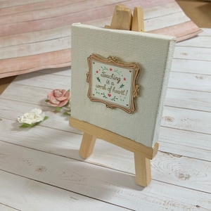 May include: A small white canvas with a gold framed pin that reads "Teaching is a work of heart!" The canvas is on a small wooden easel.