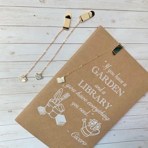 May include: Two gold bookmark chains with decorative charms. One chain has a teapot charm and the other has a book charm. The chains are attached to a brown notebook with the quote "If you have a garden and a library you have everything you need." - Cicero.