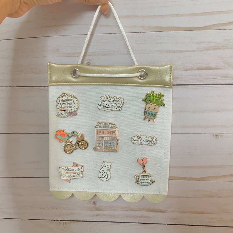 May include: A white canvas bag with a gold top and scalloped trim. The bag has a white cord handle and is decorated with various enamel pins featuring whimsical designs, including a tea shop, a cat, a potted plant, and a heart.