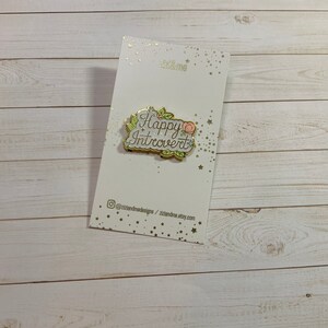 May include: Gold enamel pin with a white background and green leaves. The pin says "Happy Introvert" in gold lettering with a pink flower.