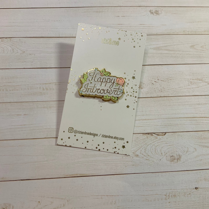 May include: Gold enamel pin with a white background and green leaves. The pin says "Happy Introvert" in gold lettering with a pink flower.