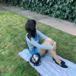 May include: A young person wearing a gray long-sleeved shirt and denim overalls sits on a pink and white plaid blanket. They are wearing black slip-on shoes and a black heart-shaped backpack with colorful patches.