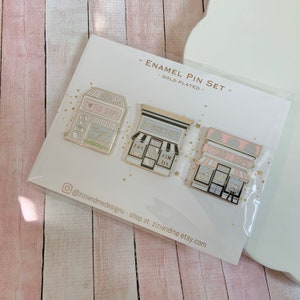May include: A gold-plated enamel pin set featuring three storefront designs: a tea shop labelled I HEART TEA SHOPS, a bookshop labelled I HEART BOOKSHOPS, and a craft shop labelled I HEART CRAFT SHOPS. The pins are mounted on a white display card.