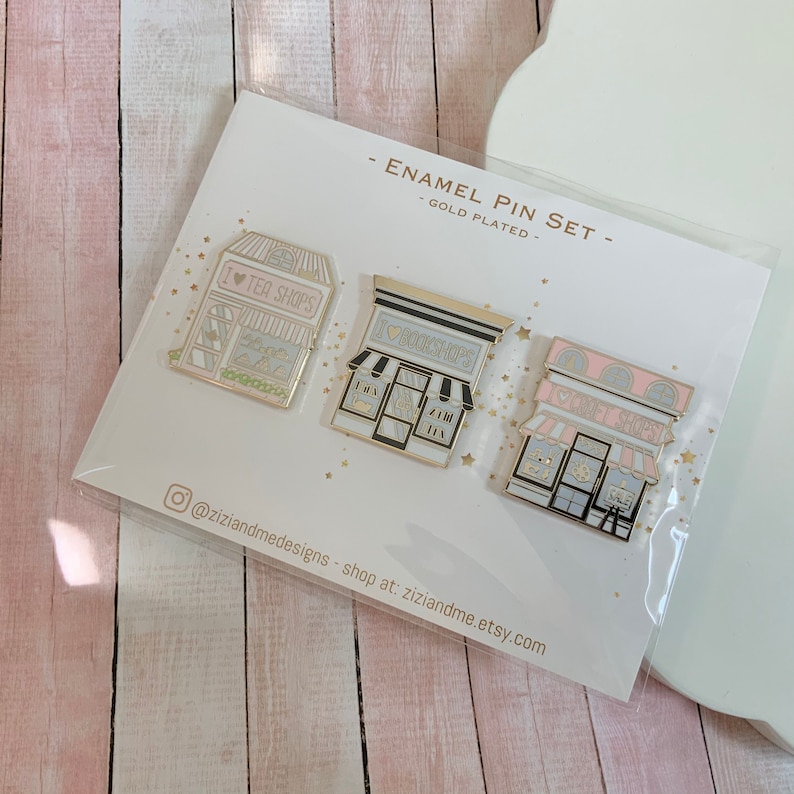 May include: A gold-plated enamel pin set featuring three storefront designs: a tea shop labelled I HEART TEA SHOPS, a bookshop labelled I HEART BOOKSHOPS, and a craft shop labelled I HEART CRAFT SHOPS. The pins are mounted on a white display card.