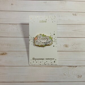 May include: A gold-colored enamel pin with a white background and a floral design. The pin says "Happy Extrovert" in pink and black lettering.