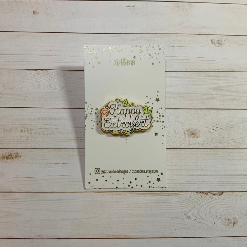 May include: A gold-colored enamel pin with a white background and a floral design. The pin says "Happy Extrovert" in pink and black lettering.