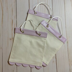 May include: Three white fabric bags with lavender colored leather trim. The bags have scalloped edges and are lined up in a row. The bags are on a light wood surface.