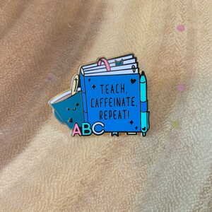 May include: A blue enamel pin with a closed book, a coffee cup, and a pen. The book has the text "Teach, Caffeinate, Repeat!" and the letters "ABC" are below the book.