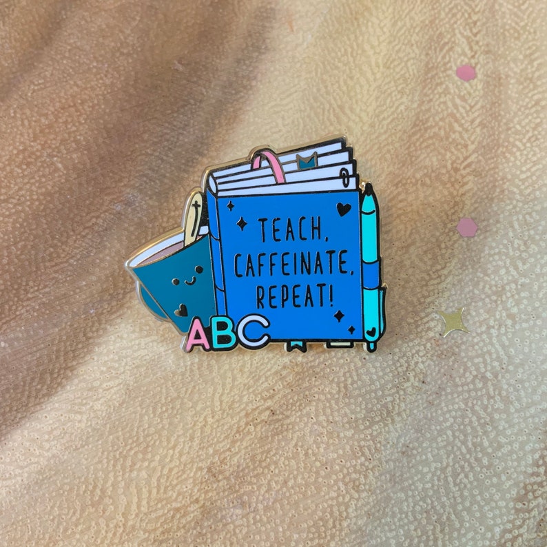 May include: A blue enamel pin with a closed book, a coffee cup, and a pen. The book has the text "Teach, Caffeinate, Repeat!" and the letters "ABC" are below the book.