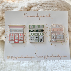 May include: A set of three enamel pins on a white display card labelled Enamel pin set. The pins depict storefronts with the text I Love Bookshops, I Love Craft Shops, and I Love Tea Shops. The card includes the social media handle @ziziandmedesigns.