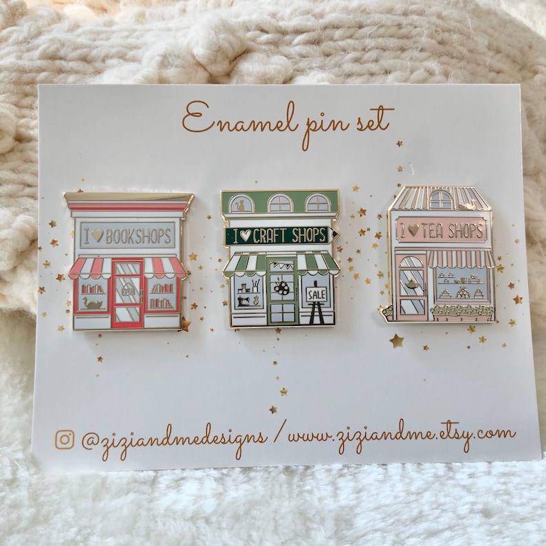 May include: A set of three enamel pins on a white display card labelled Enamel pin set. The pins depict storefronts with the text I Love Bookshops, I Love Craft Shops, and I Love Tea Shops. The card includes the social media handle @ziziandmedesigns.
