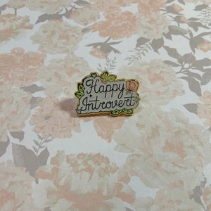 May include: A gold-colored enamel pin with a white background and green and pink floral accents. The pin says "Happy Introvert" in cursive writing.