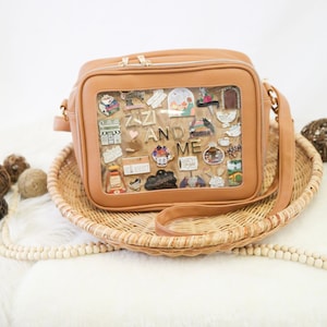 May include: A tan faux leather crossbody bag with a clear plastic window on the front. The window is filled with colourful pins and the text "Zizi and Me" is spelled out with gold letters.