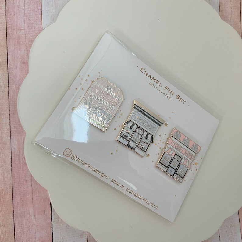 May include: A set of three gold-plated enamel pins featuring storefront designs labelled I LOVE TEA SHOPS, I LOVE BOOKSHOPS, and I LOVE CRAFT SHOPS. The pins are displayed on a white backing card with the text ENAMEL PIN SET and gold star accents.