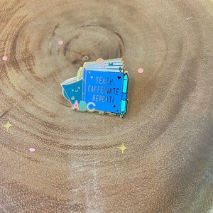 May include: Gold enamel pin featuring a blue notebook with the text "Teach. Caffeinate. Repeat!" and a coffee cup with the letters "ABC" in pink, yellow, and blue.