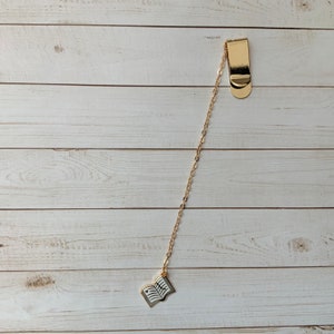 May include: Gold bookmark with a chain and a book charm. The book charm is open and has a white background with black lines.