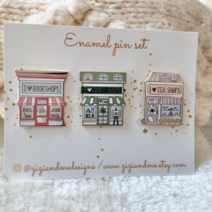 May include: A set of three enamel pins on a white display card labelled Enamel pin set. The pins depict storefronts with the text I heart BOOKSHOPS, I heart CRAFT SHOPS, and I heart TEA SHOPS. The card includes the text @ziziandmedesigns / www.ziziandme.etsy.com.