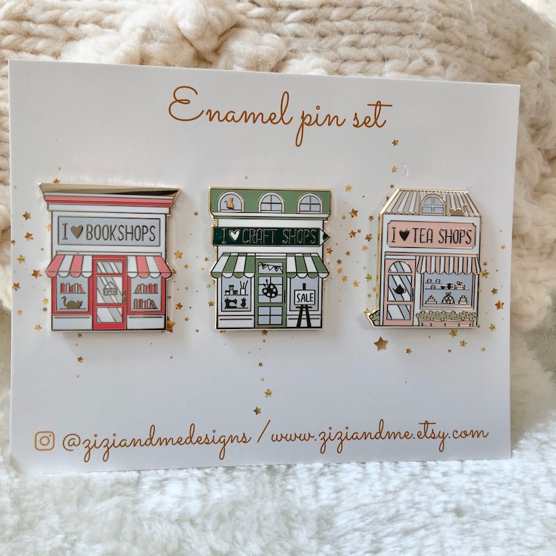 May include: A set of three enamel pins on a white display card labelled Enamel pin set. The pins depict storefronts with the text I heart BOOKSHOPS, I heart CRAFT SHOPS, and I heart TEA SHOPS. The card includes the text @ziziandmedesigns / www.ziziandme.etsy.com.