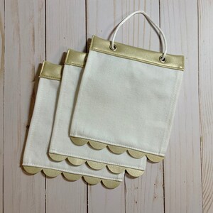 May include: Three white canvas bags with gold scalloped edges and gold faux leather trim. The bags have a gold grommet and a white string handle.