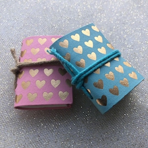 May include: Two small notebooks with gold heart patterns on the covers. One notebook is pink and the other is blue. Both notebooks are tied with twine.