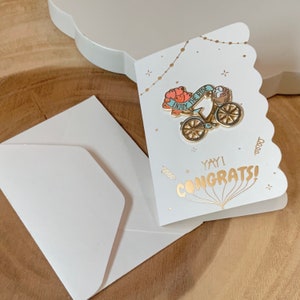 May include: A white card with gold lettering that says "Yay! Congrats!" and a gold pin of a bicycle with the words "Enjoy the Ride" on a banner. The card has a scalloped edge and is decorated with gold stars and dots.
