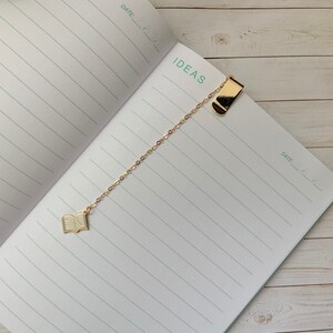 May include: A gold chain bookmark with a book charm and a clip at the top. The bookmark is on a lined notebook with the word "IDEAS" written at the top.