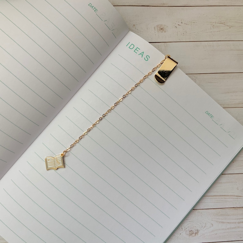 May include: A gold chain bookmark with a book charm and a clip at the top. The bookmark is on a lined notebook with the word "IDEAS" written at the top.