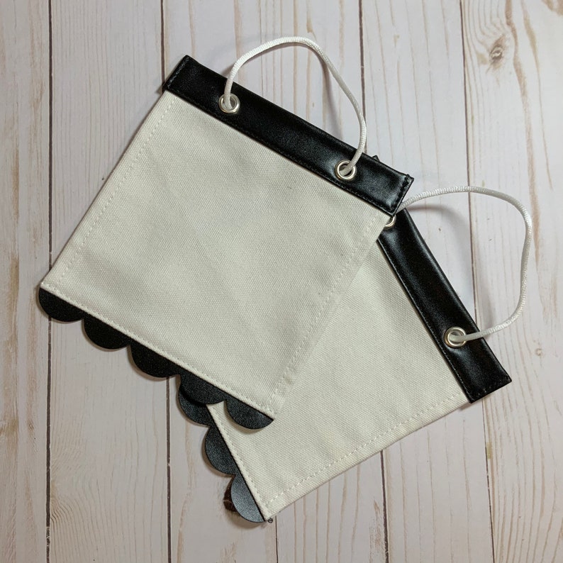 May include: Two white fabric banners with black scalloped edges and black leather trim. Each banner has two white eyelets at the top for hanging.