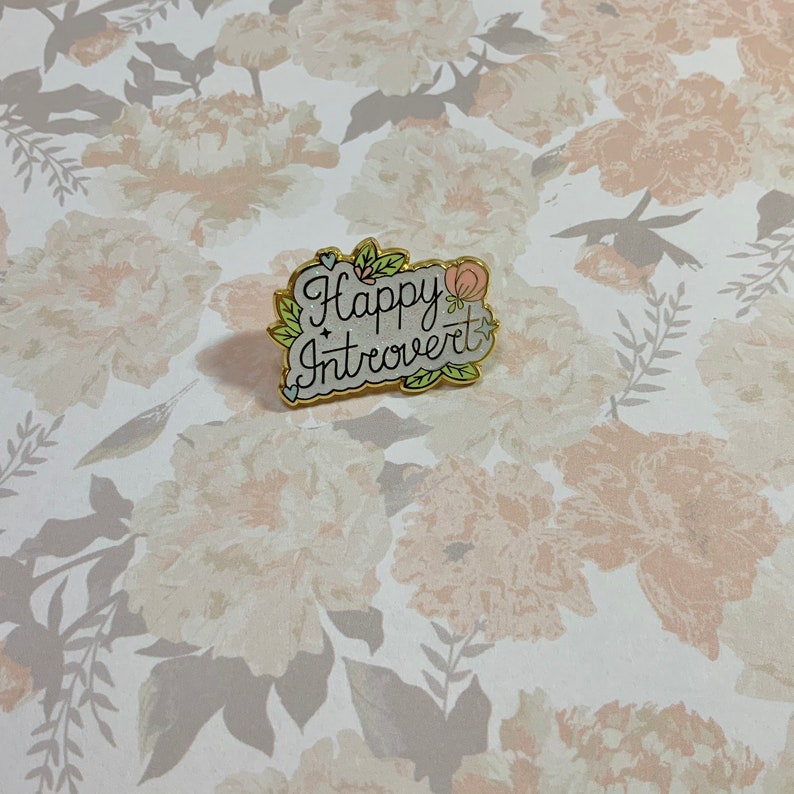 May include: A gold enamel pin with a white background and the text "Happy Introvert" in black script. The pin is decorated with small pink and green flowers.