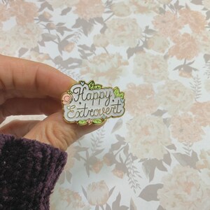 May include: White enamel pin with gold trim featuring the text "Happy Extrovert" surrounded by pink and green floral accents.