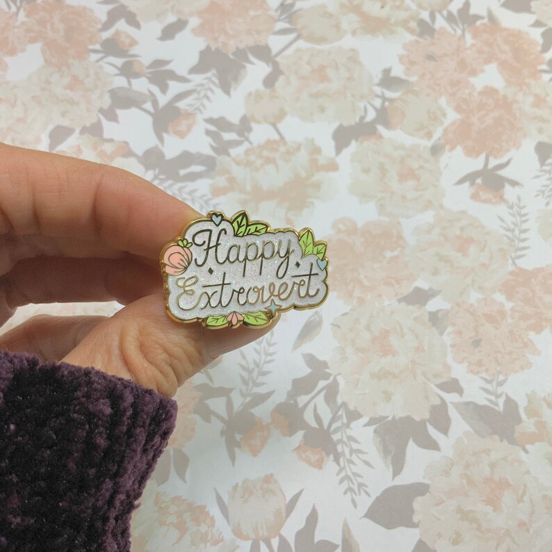 May include: White enamel pin with gold trim featuring the text "Happy Extrovert" surrounded by pink and green floral accents.