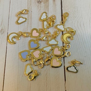 Könnte beinhalten: Eine Sammlung von goldenen Charms in verschiedenen Formen und Farben, darunter Herzen, Wolken, Monde und Sterne. Die Charms sind an einer goldenen Kette befestigt.