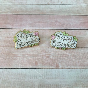 May include: Two gold enamel pins with white backgrounds and floral accents. The left pin says "Happy Introvert" and the right pin says "Happy Extrovert".