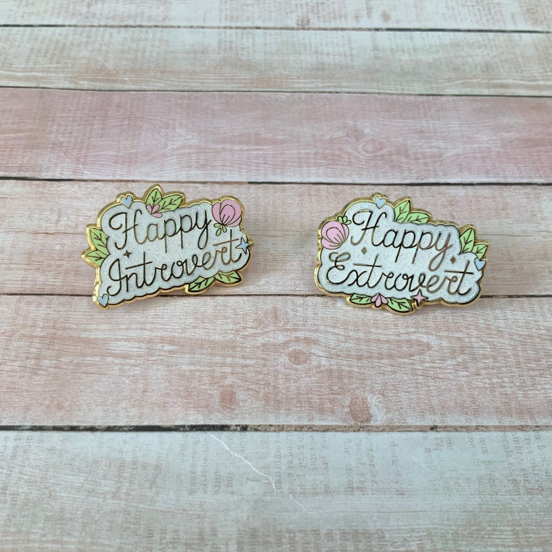 May include: Two gold enamel pins with white backgrounds and floral accents. The left pin says "Happy Introvert" and the right pin says "Happy Extrovert".