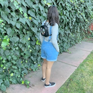 May include: A person wearing a denim overall dress with a black backpack with colorful patches. The person is standing in front of a green ivy wall.