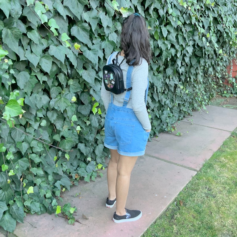 May include: A person wearing a denim overall dress with a black backpack with colorful patches. The person is standing in front of a green ivy wall.