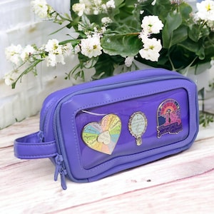 Peut inclure: Une trousse à crayons violette avec une fenêtre transparente montrant trois épingles en émail. Les épingles sont un cœur arc-en-ciel avec les mots "Embracing", "Organizing", "Friends", "Creativity", "Laughter", "Kindness", "Joy", "Love", "Hope", "Faith", et "Trust" autour du cœur, un miroir doré avec les mots "The beauty you see in yourself is reflected in the world around you", et une épingle bleue et rose avec un château et les mots "Dream Big, Little One".