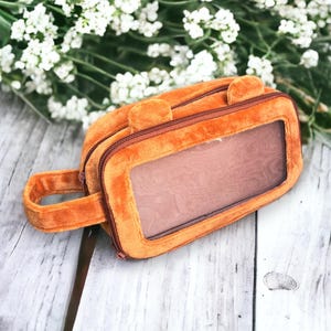 May include: A brown plush pencil case with a clear plastic window and a zipper closure. The case has a handle and two fuzzy ears on the top.