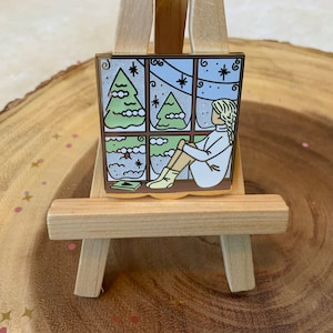 May include: A gold-toned enamel pin featuring a window scene with a person sitting by the window, looking out at two snow-covered pine trees. The window is divided into four panes and has a white frame. The person is wearing a white sweater and blue pants. The background is a light blue sky with white snowflakes.