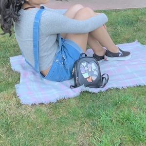 May include: A person wearing a grey sweater and denim overalls sits on a pink and white plaid blanket with a black heart-shaped backpack with a clear front pocket. The backpack has various colorful stickers on the front.
