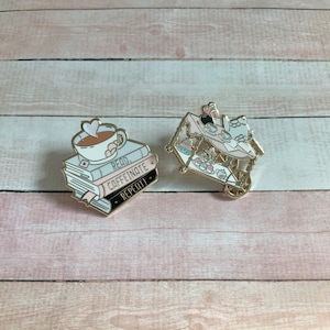 May include: Two enamel pins. One pin shows a cup of coffee with a heart on top of a stack of books. The books read "Read, Caffeinate, Repeat!". The other pin is a pink and gold tea cart with desserts.