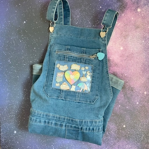 May include: Blue denim overalls with a clear plastic pocket on the front. The pocket is filled with colorful pins and patches, including a rainbow heart, a coffee cup, and a cat.