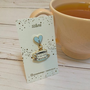 May include: A gold-colored enamel pin featuring a teacup design with the text "Op-tea-mist!" and a light blue heart. The pin is attached to a white card with gold star accents and the text "zizi&me". A peach-colored mug filled with tea is in the background.