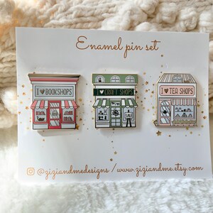 May include: A set of three enamel pins on a white display card labelled Enamel pin set. The pins depict storefronts with the text I heart bookshops, I heart craft shops, and I heart tea shops. The card includes the URL ziziandme.etsy.com.