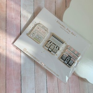 May include: A set of three gold-plated enamel pins on a display card labelled ENAMEL PIN SET. The pins feature storefront illustrations with the text I LOVE TEA SHOPS, I LOVE BOOKSHOPS, and I LOVE CRAFT SHOPS, each with distinct pastel colour accents.