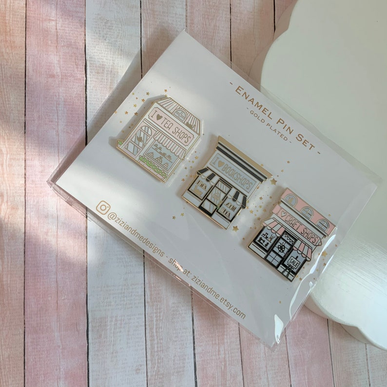May include: A set of three gold-plated enamel pins on a display card labelled ENAMEL PIN SET. The pins feature storefront illustrations with the text I LOVE TEA SHOPS, I LOVE BOOKSHOPS, and I LOVE CRAFT SHOPS, each with distinct pastel colour accents.