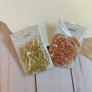 Könnte beinhalten: Zwei Packungen mit goldenen und roségoldenen Büroklammern mit dem Text "zizi&me" auf der Vorderseite jeder Packung.